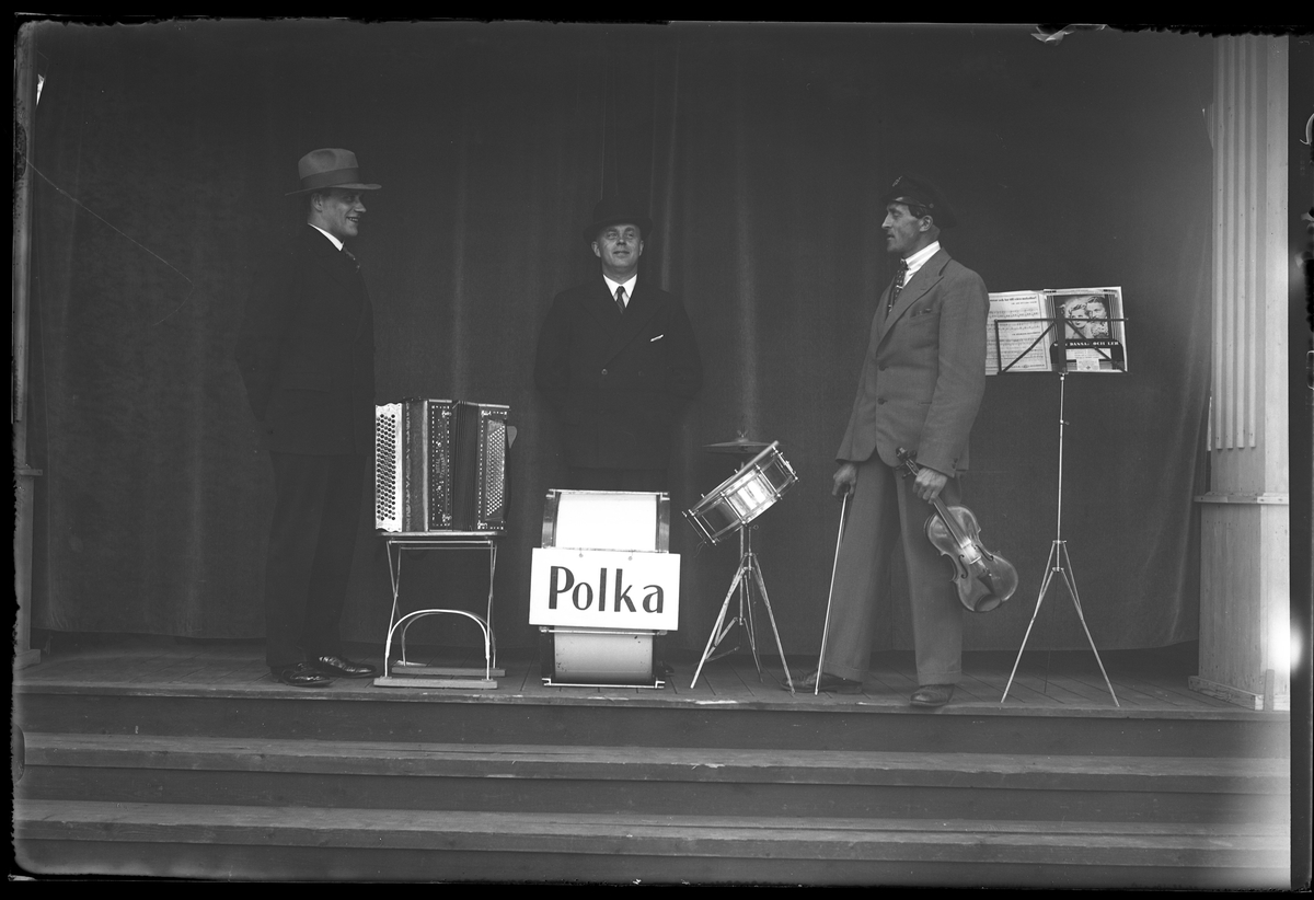 Tre män står på en scen. Den ena står bredvid ett dragspel, en annan vid två trummor och den tredje har en fiol i handen. I fotografens anteckningar står det "Gammalmusiken i Parken".