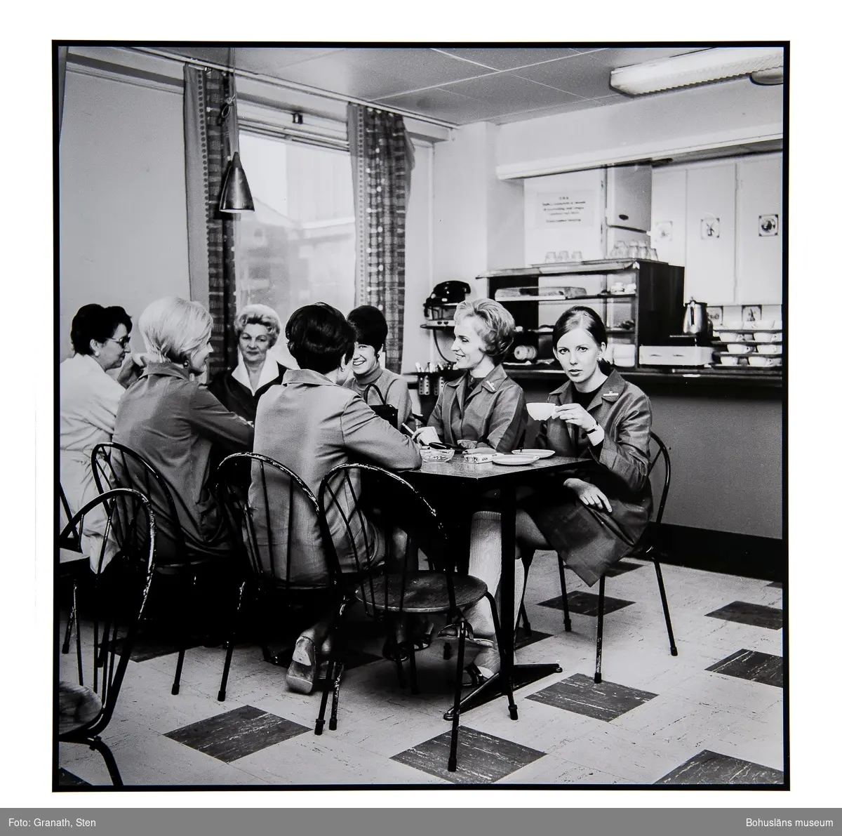Fikapaus på EPA-varuhuset. Sju kvinnor sitter kring ett rektangulärt bord. Fyra av kvinnorna har likadana arbetsuniformer på sig. Bakom bordet finns en disk med porslin, en kylmonter och en kaffeperkulator.