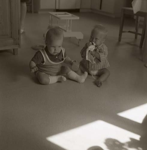 Två bebisar sitter på golvet i ett rum och den ena leker med en leksak. Framför dem kastas solkatter in genom fönstret, 1972.