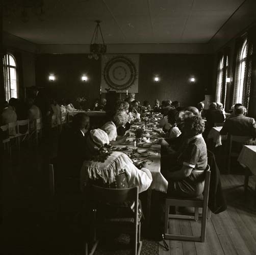 Ett stort sällskap är församlade runt några långbord i en stor lokal,1983.