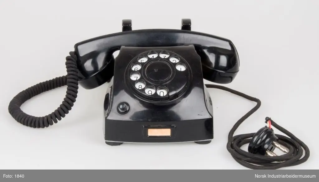 Telefon med drejeskive af sort bakelit. Sort ledning med tekstilisolering. Telefonen bærer præg af brug. Den blanke bakelitoverflade er slidt mat en del steder.