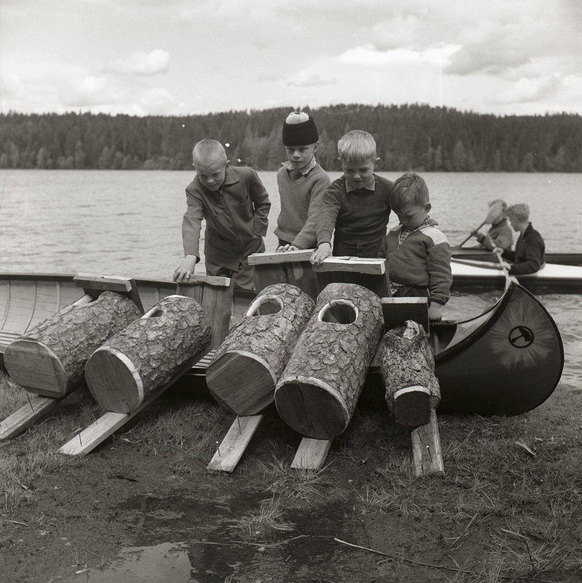 Några barn står bakom en kanot och tittar på några fågelholkarna som ligger på marken, Hagberget  den 10 maj 1959.
