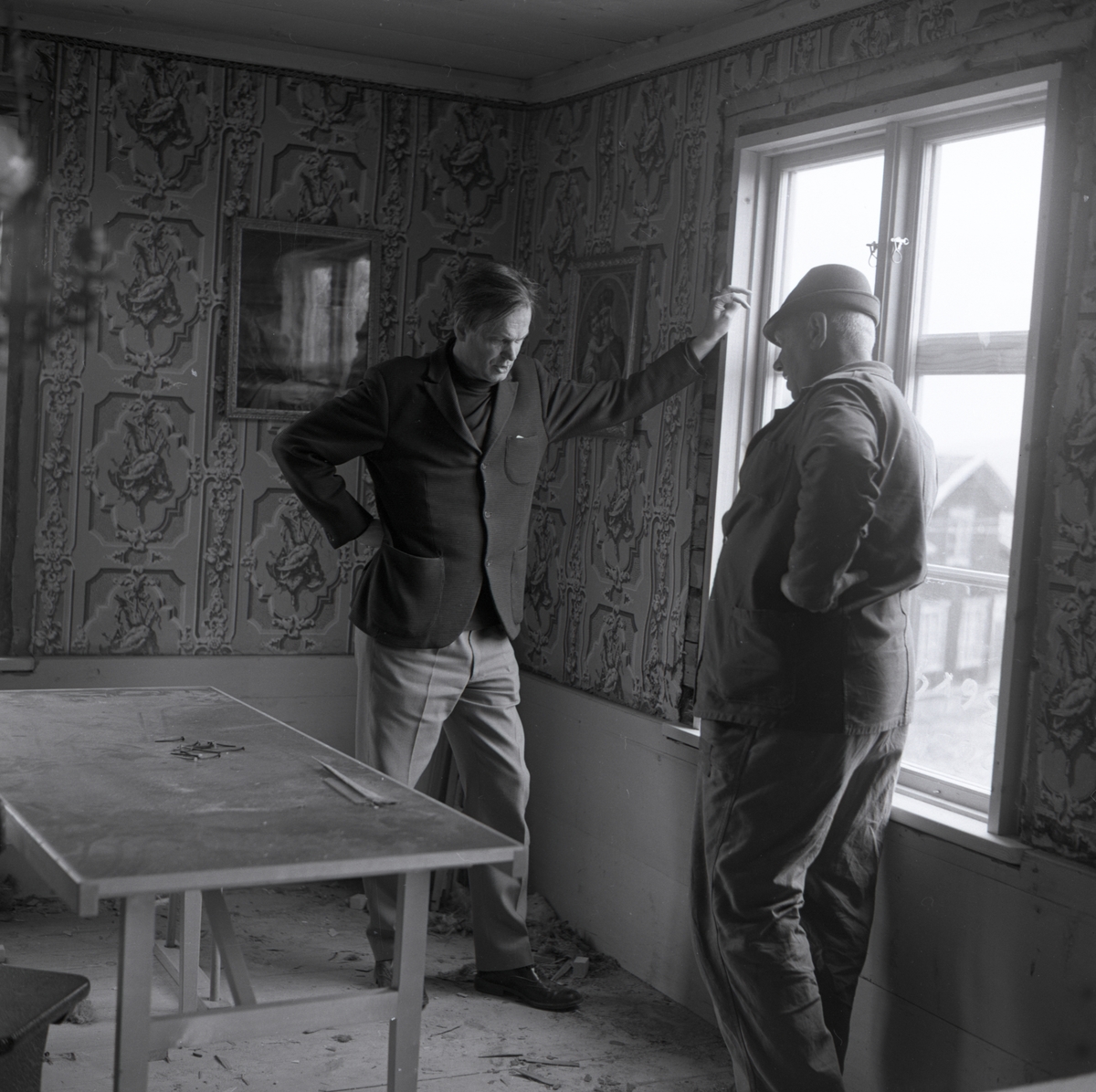 I ett av rummen vid hembygdsgården i Rengsjö har en renovering påbörjats. Två män diskuterar stående intill ett fönster, 1968.