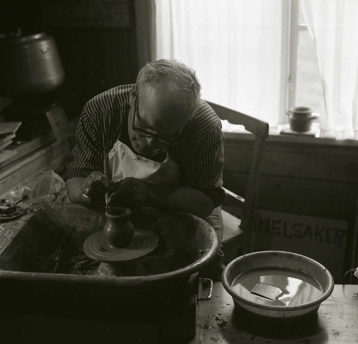 En man sitter vid en drejskiva och drejar ett kärl av lera med hjälp av ett verktyg under Gammelvärldens dag vid Hembygdsbyn 1980 . Mannen har ett förkläde och glasögon på sig. Bakom honom syns ett fönster med gardin och en kruka på fönsterkarmen.