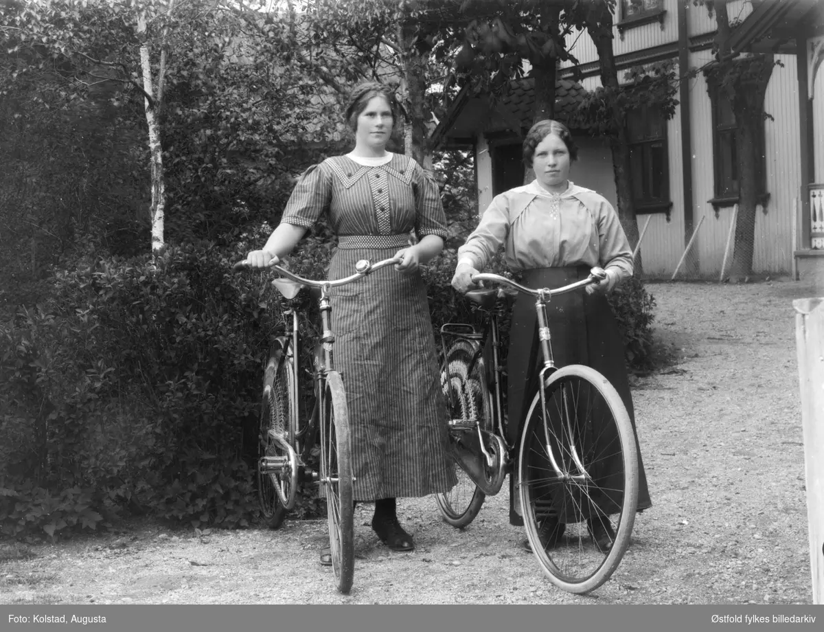 To unge kvinner med sykkel  på tunet utenfor våningshus. Begge kvinnene er ukjente.