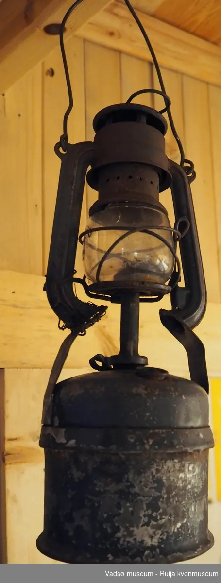 Rusten stormlykt. Sylinderformet parafinbeholder med brenner på toppen. Bobleformet brennerrør i glass. Ramme i metall med jernhank på toppen.