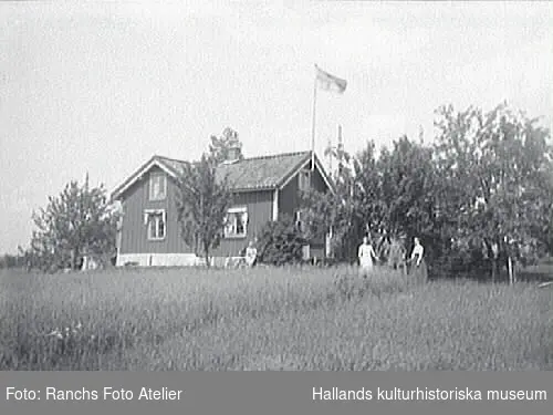 Villa i Haby, Örby. A. Alm är troligen ägare till huset. Vid husgaveln är en flaggstång fästad och det kan vara finska flaggan som är hissad. Fem personer uppehåller sig vid huset, bland annat en flicka med cykel. I förgrunden är en havreåker.
