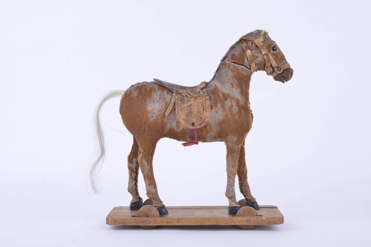 Modell av hest montert på en rektangulær treplate med fire hjul av tre. Hesten er utformet av skinn og fyllt med spon. På utsiden av skinnet er det limt på imitert hår/tekstil. Sal og bissel av lær.