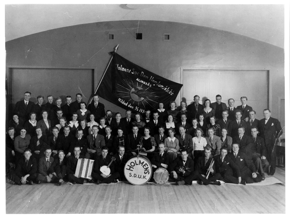 Gruppbild av ett 70-tal kvinnor och män med en stor flagga i bakgrunden med texten " Holmens Soc Dem Ungdomsklubb, Alingsås, Bildad den 3/9 1934". 6 av männen i främsta raden sitter med var sitt musikinstrument.