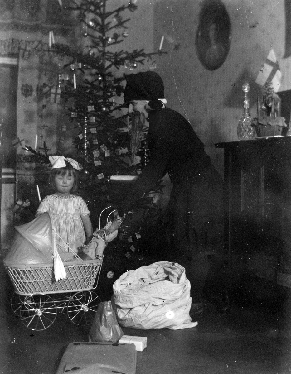 En jultomte står framför en julgran och håller i en dockvagn med en docka i, ett barn står jämsides och blundar.
