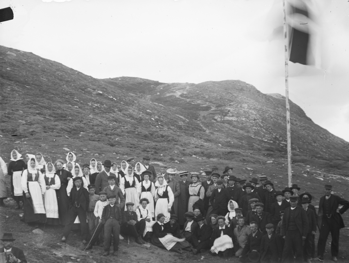 Stor gruppe festkledde folk, mange med bunad, står ute i fjellandskap ved flaggstang. Antatt ved Bessheim