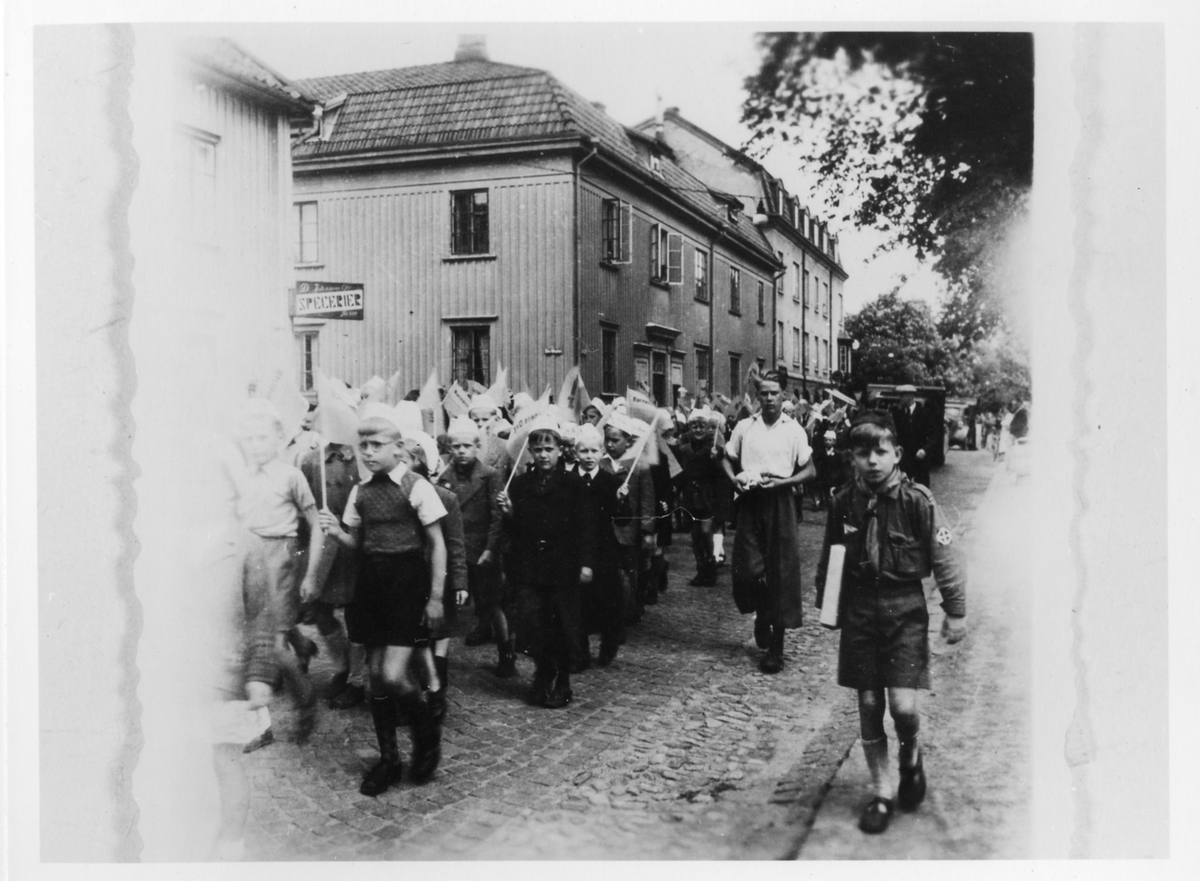 Långt och brett led av scouter, barnen går nerför Södra Ringgatan med vita mössor och var sin vit rektangulär flagga i hand. Till höger en pojke i full scoutmundering bärande på en bok under högra armen. 
Kvarteret Cederberg till vänster och Östra Vattugränd in till vänster.