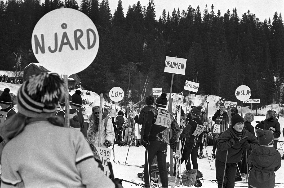 Ungdom med ski samlet rundt mange plakater med forskjellige idrettslag, "Aprilspøken" slalåmrenn i Tryvannskleiva. Fotografert 23. mars 1969.