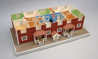 Modell i skala 1:50 av falurött tvåvåningshus med valmat sadeltak. Stående plank, tegeltak, och vita skorstenar. Byggnaden innehåller fyra lägenheter om 4 rum och kök. Inredning saknas.