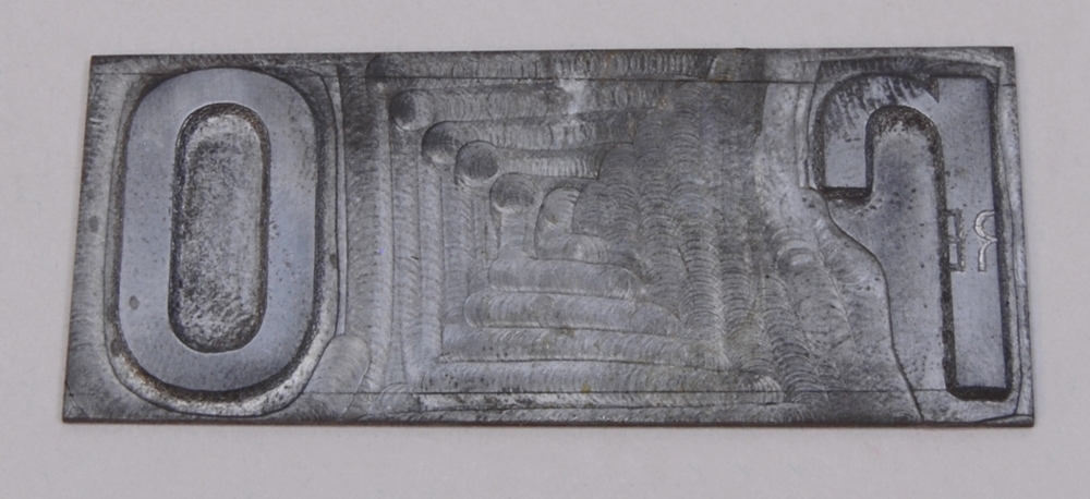 Rektangulär kliché av silverfärgad metall. På höger sida finns bokstaven "r" i relief, och på vänster sida finns bokstaven "o" i relief.
