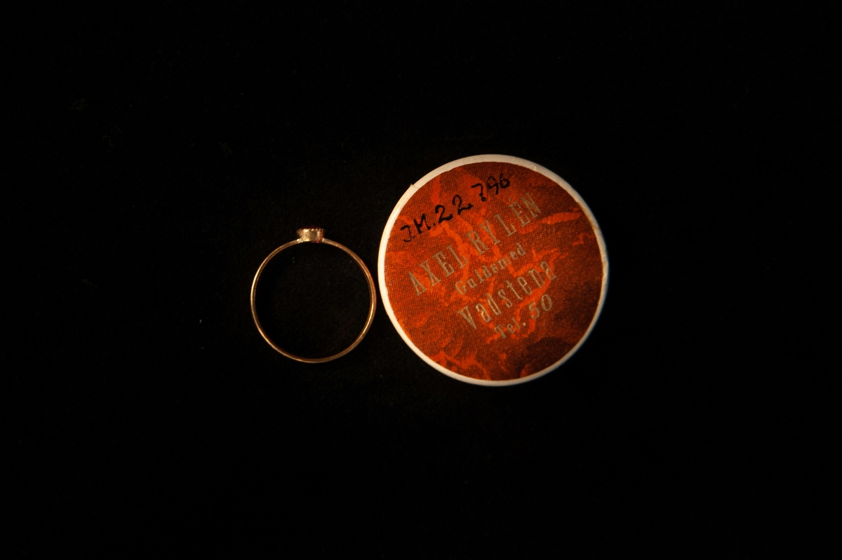 Smal guldring med infattad ljusröd sten (troligen simili (glas), möjligen rubin(?). En tillhörande rund smyckesask i papp.
