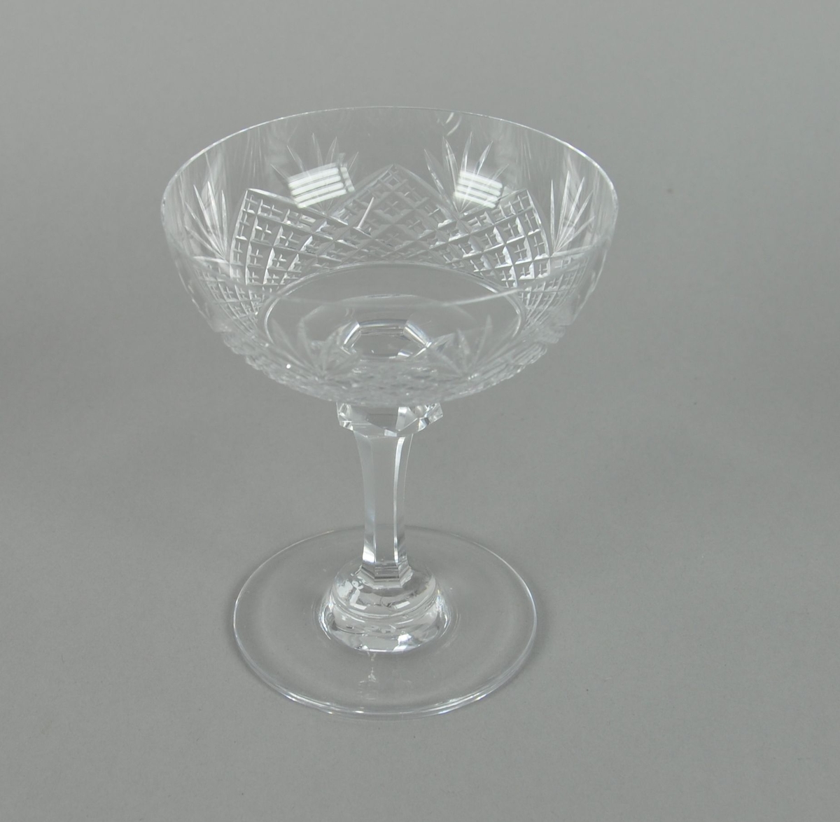 Sjampanjeglass av krystall. Glasset har rund form med sekskantet stett og rund sokkel. På utsiden av glasset er det innrisset dekor av kryssmønstre og palmetter.