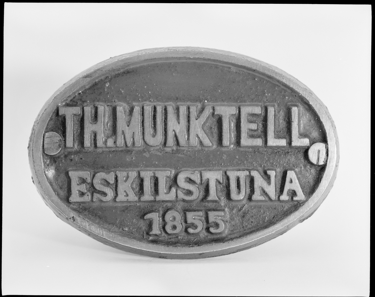 Oval skylt i mässing med text i relief på rödmålad botten:
"TH.MUNKTELL
  ESKILSTUNA
      1855".
Från loket Fryckstad.