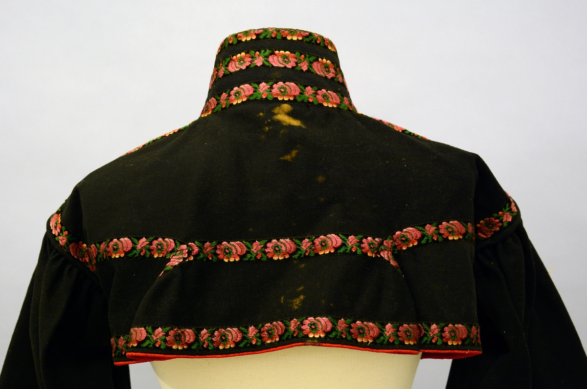 Kvinnetrøye av svart klede, kanta med kjøpeborder i mønster av raudt og grønt på svart botn. Høg krage, ope bryst. Fra protokoll.