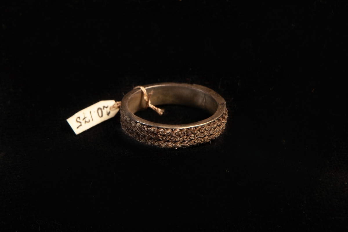 En ring av förgyllt silver. Fattat i ringen en dubbelflätad mörkbrun
hårfläta. Front i guld med ingraverade ägarinitialer: "J.E.W." Mycket otydliga stämplar invändigt.