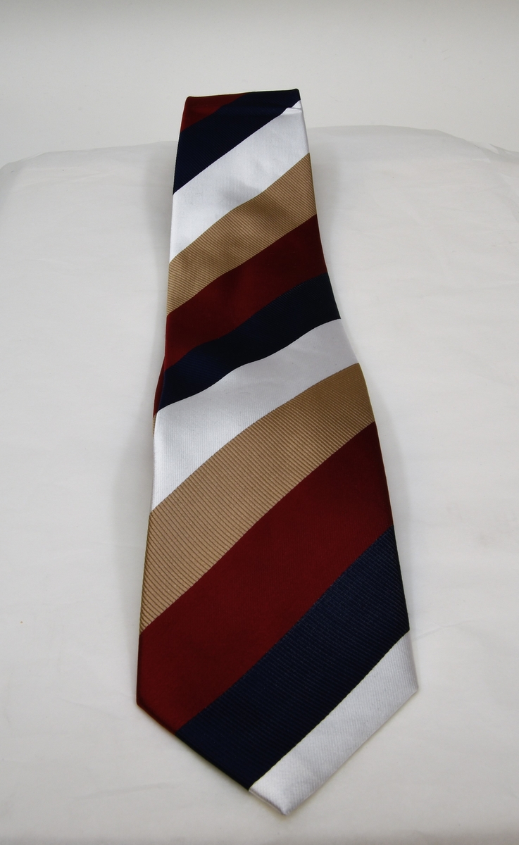 Slips av silke med diagonalrandigt mönster i färgerna vit, blå, röd och ljusbrunt. På baksidan av slipsen sitter det två lappar.