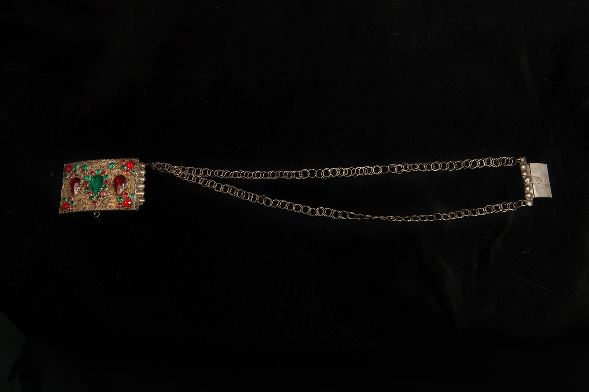 Ett halslås av silver med två enkla kedjor. Fronten prydd med filigranarbete med infattade, slipade glasstenar i rött och grönt. Undertill liten krok för hjärtformat hänge eller dyllikt.