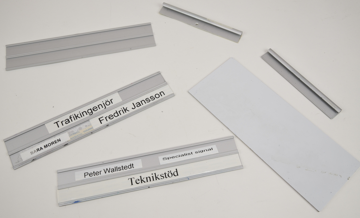Namnskyltshållare av obehandlad extruderad aluminium (:1-3) med tillhörande väggfästen (:4-5) och en aluminiumplatta med vit yta som fungerar som en mini-whiteboard (:6). Skylthållarna har plats för två rader textremsor som skjuts in från sidan. En av hållarna saknar namn men två av dem har både namn och titlar med svart text på vita Dymo-remsor. Remsorna med "Teknikstöd" och "Fredrik Jansson" är inte fastklistrade utan har kvar Dymo-remsans täckpapper.