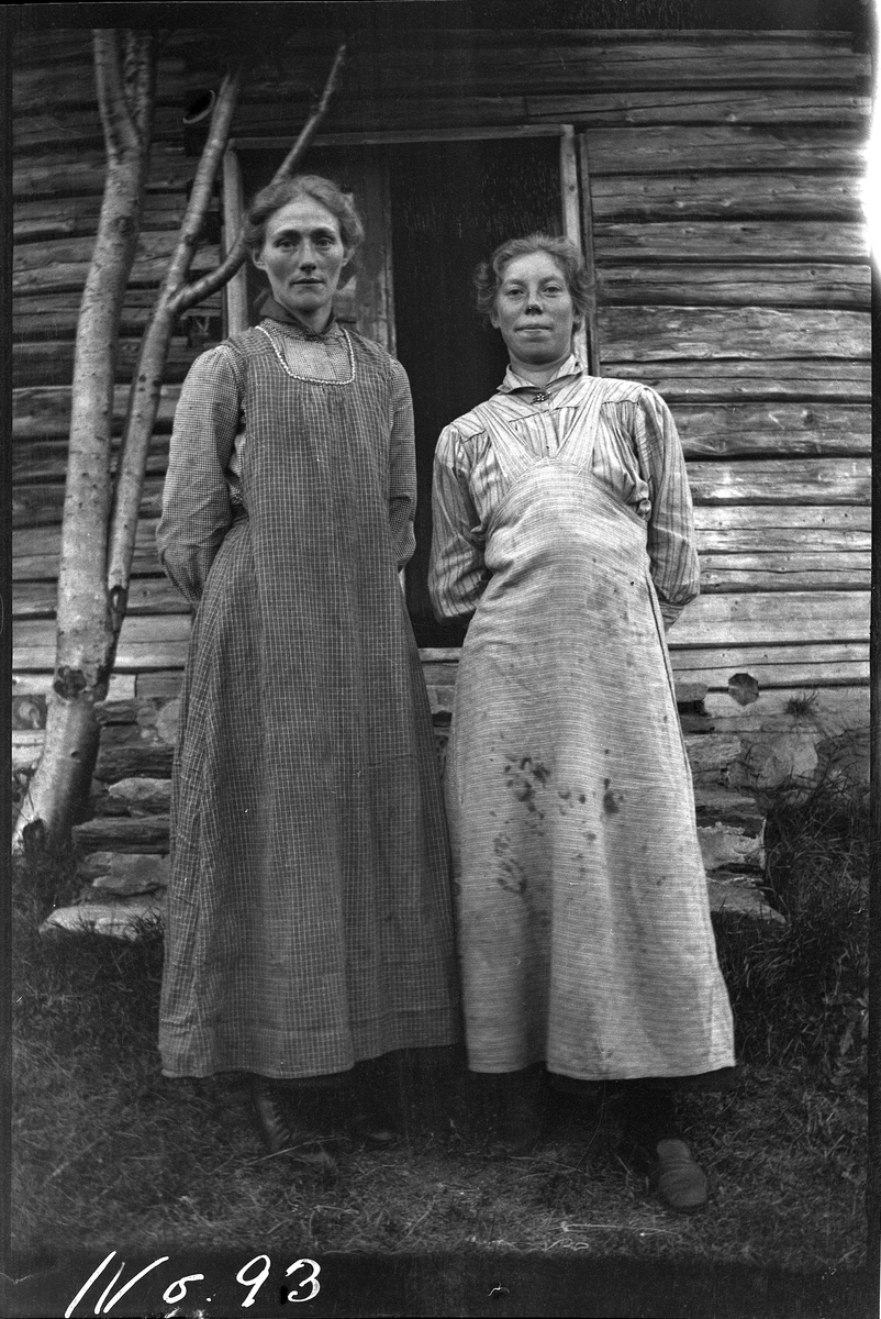Portrett av to kvinner, tatt stående foran en laftet bygning.