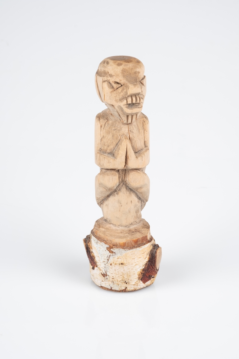 Figur skåret ut av en stubbe. Figuren kan minne om skulpturer fra Søramerikanske stammer. Figuren er av et menneske som sitter på en stubbe med håndlfatene slått sammen.