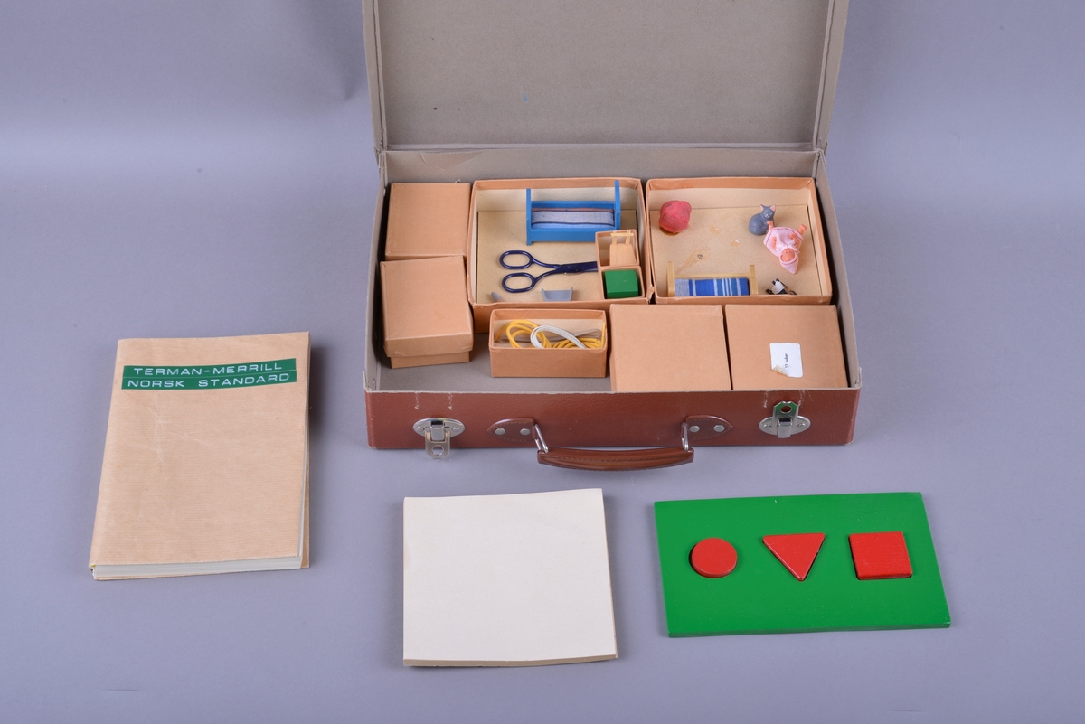 Rektangulær brun koffert av papp, med håndtak og lås. Inne i kofferten er det små pappesker. Pappeskene inneholder små treklosser, plater med pålimte miniatyr-leker. Blant annet dukke, hund seng, saks, pipe. I tillegg er det flere små knapper, notatblokk og tekstilbånd. I kofferten ligger også en bok merket "TERMAN-MERRILL // NORSK STANDARD".