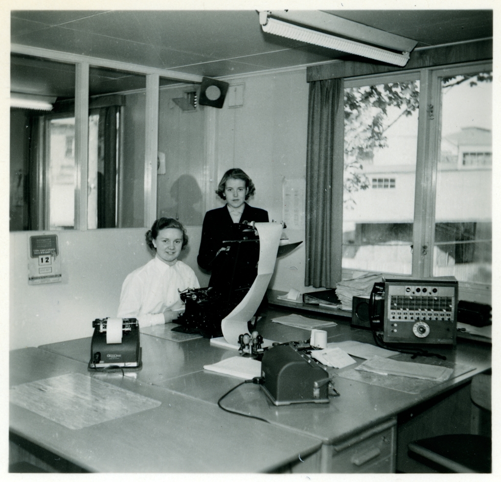 Två av personerna som arbetade på Monarks kontor. Den ena kvinnan sitter ner vid en skrivmaskin(?) och till höger om henne står Gunnel Andersson. På bordet står också en telefonväxel.