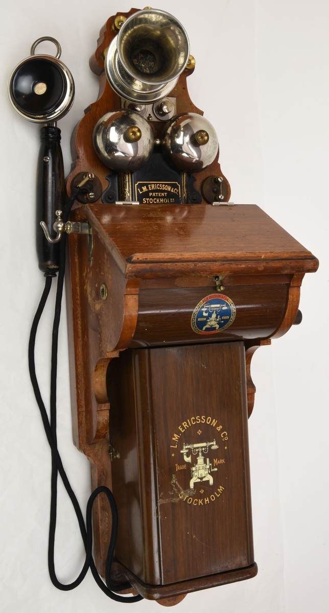 En väggtelefon av trä från tidigt 1900-tal gjord av L.M. Ericsson.

Mikrofonen är monterad direkt på telefonen och ser ut som en metalltratt, hörapparaten är handhållen och är kopplad till telefonen via en elkabel. På väggtelefonens överdel finns skruvbara mässingsfästen. Nedanför mikrofonen finns en ringklocka och ett svart påskruvat metallhölje med påmålad ornamentik. Mitt på väggtelefonen sitter en utstickande trälåda som innehåller mekaniska komponenter som rör på sig när man rör på den vev som sticker ut ur lådans högra sida. På undersidan av locket till trälådan finns en pappersetikett med en förteckning över telefonen. Nedanför trälådan finns ett hyllplan som täcks av en metallkåpa (med målad träimitation), i det utrymmet ska det finns ett batteri.