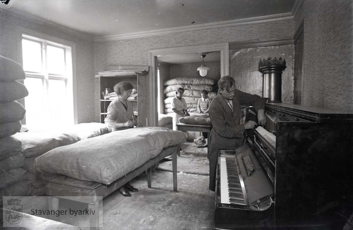 Pianoet stemmes. Noen kvinner jobber med madrasser eller dyner som ligger stablet opp langs veggen.
