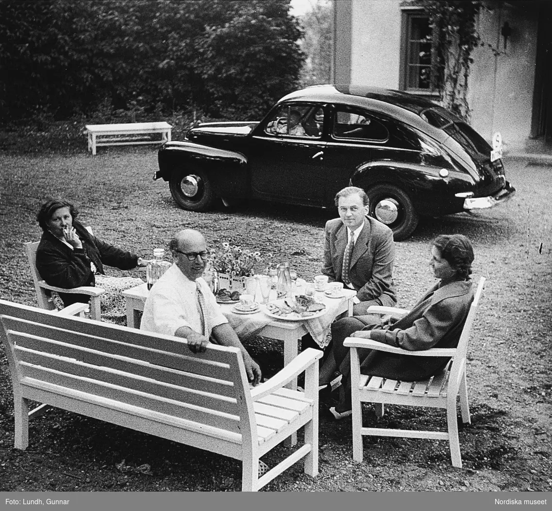 Personer sitter kring ett dukat bord på trädgårdsmöbler i en trädgård. I bakgrunden en Volvo PV årsmodell 1947.