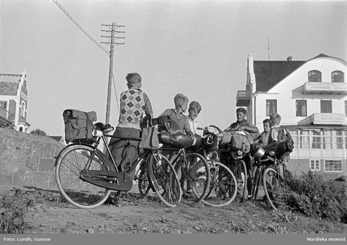 En grupp pojkar med cyklar står vid vägen utanför Grand Hotell i  Mölle ( i bakgrunden t.h.), Cyklarna är lastade med ryggsäckar och sovsäckar,