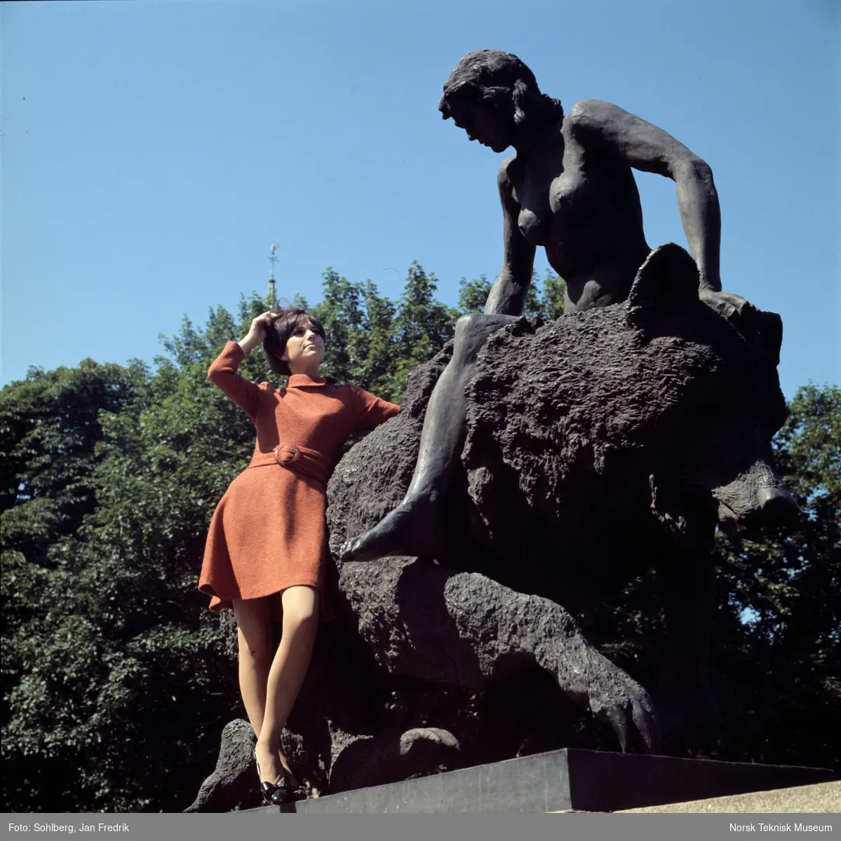 Motebilde i farger. Kvinnelig modell i rødbrun ullkjole av norsk fabrikat, fotografert foran skulpturen "Kvitebjørn kong Valemon" (1933-36) av Dyre Vaa på Ankerbrua/ Eventyrbrua i Oslo, 1968. Bildet er en del av en serie der ett av dem er publisert i ukebladet Det Nye nr. 39, 1968.