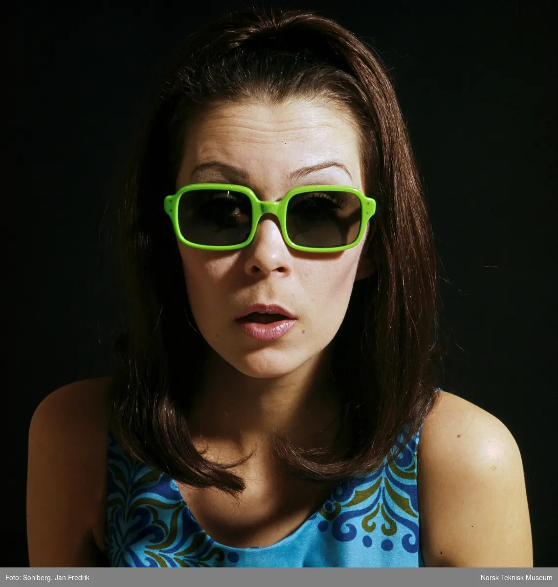 En kvinnelig modell viser fram tidenssolbrillemote. Hun har langt hår hengende løst. Hun har grønne solbriller. Hippie-stil.