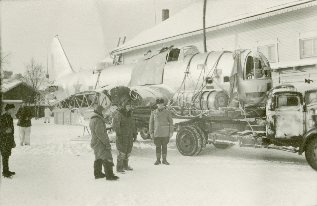 Sovjetiska flygare (i mörka overaller) som tagits som krigsfångar efter att ha skjutits ned i flygplan DB-3. Vid Svenska frivilligkåren i Finland, F 19. Flygplanet lastat på vagn efter lastbil. Militärer kring flygplanet.