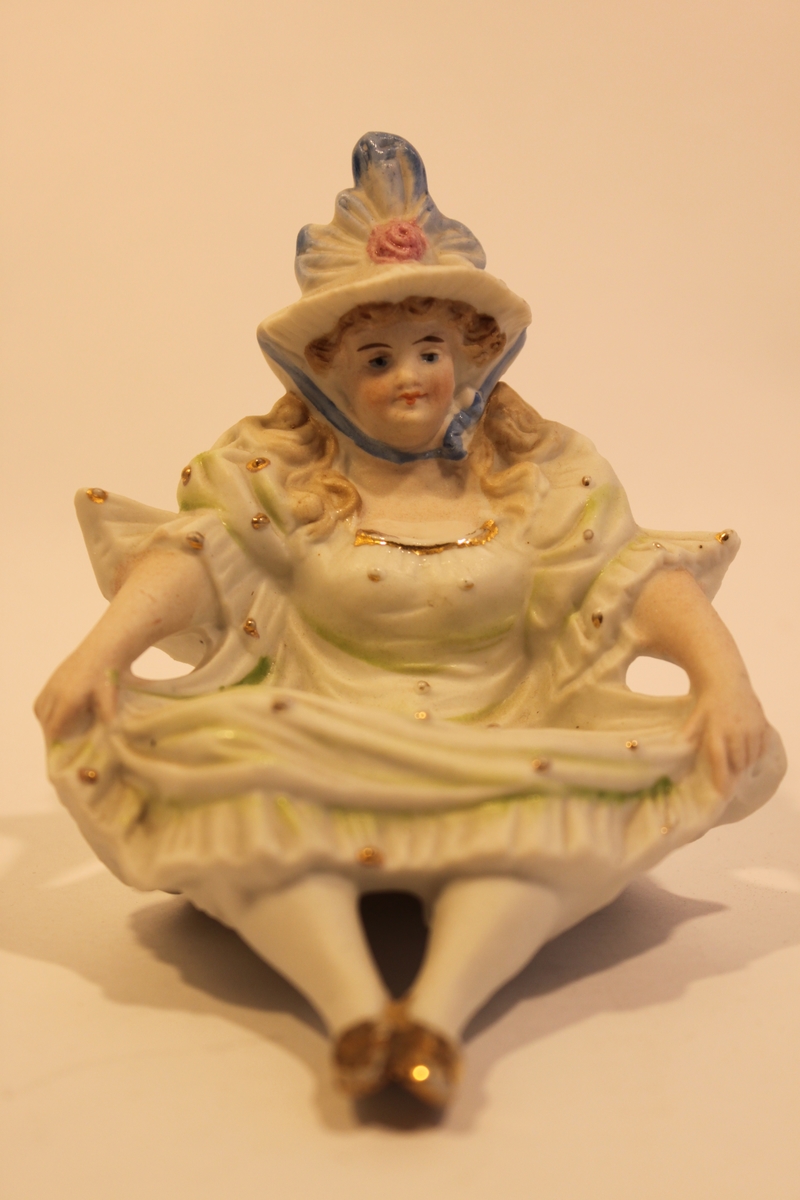 Porslinsfigur föreställande en sittande dam med hatt. Vänder man uppochner på figuren visar det sig att hon har rumpan bar.