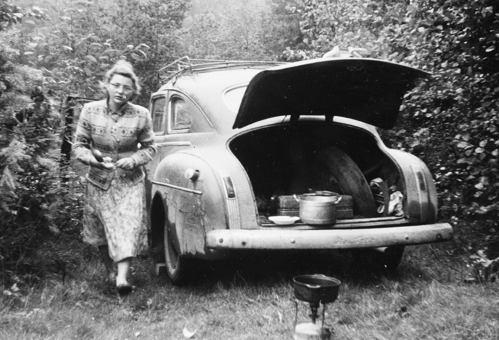 Ferietur med utleigebil i dårleg stand. Stans i Telemark. Bjørg Time (26.6.1937 - ) prøver å laga mat. I bagasjerommet kokekar og på bakken står primusen.