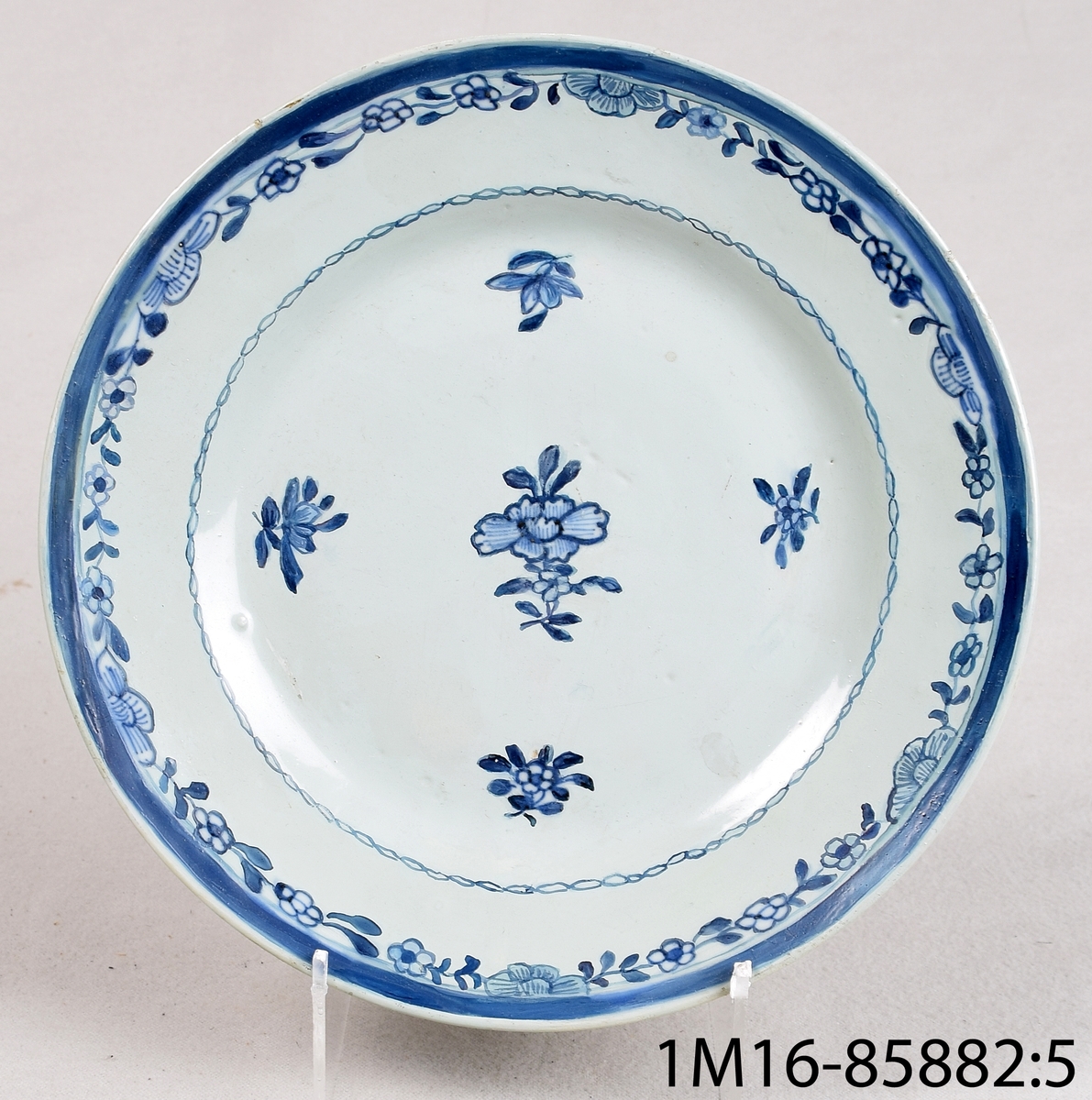 Tallrik, av blåvitt kinesisaklt porslin, flat med slät kant. Dekorerad med bård och blomma i blått.