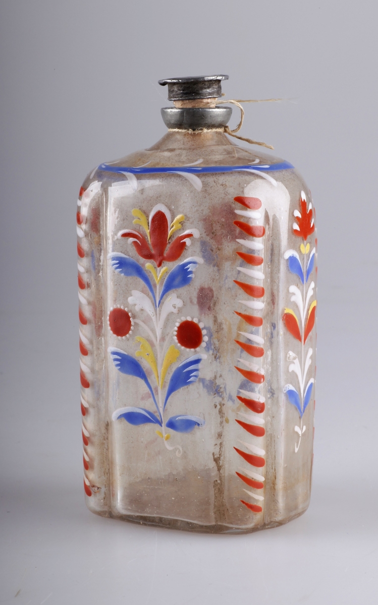 Åttekantet flaske blåst i klart glass. Med tuten innfelt i glassmassen overst, og brutt pontemerke i bunnen. Påmalt med emaljemaling eller blylakk i blått, rødt, hvitt og gult. Dekor i form av stilistiske blomster. Kork og munning i tinn. Kan være av av tysk opprinnelse i tidsepoken 1750-1850.