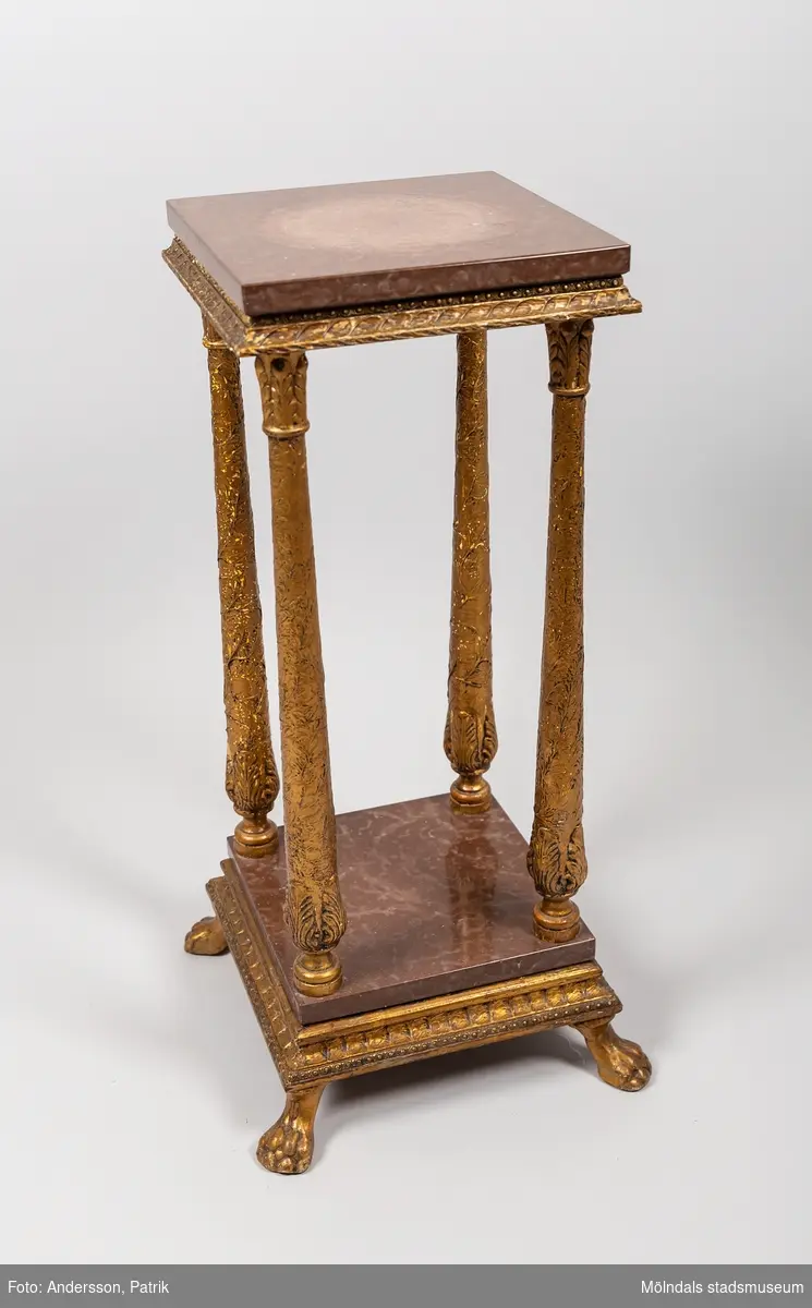 Piedestal från 1920-talet.  Piedestalen är tillverkad av förgyllt trä med rödbruna marmorskivor. Den har fyra stycken rikt utsmyckade ben och står på fyra lejontassar, som också är tillverkade av förgyllt trä.