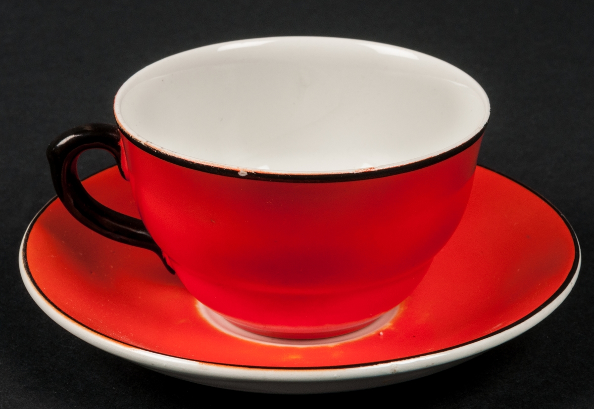Kaffekopp med fat. Röd glasyr med svarta kanter och svart hänkel. Insidan av koppen vit, samt undersidan av fatet.