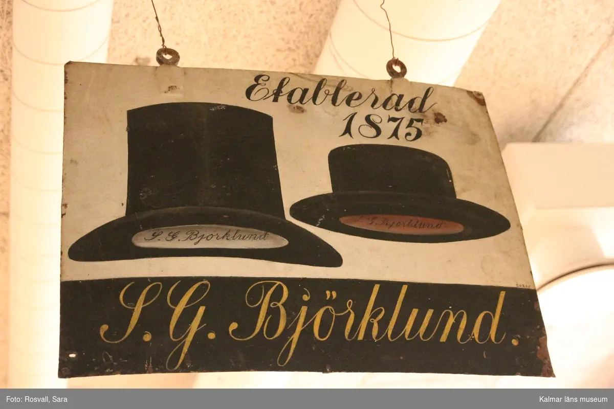 KLM 8623:1. Skylt, hattmakarskylt av plåt. Målad. Motiv med två hattar och text: Etablerad 1875 S.G. Björklund.