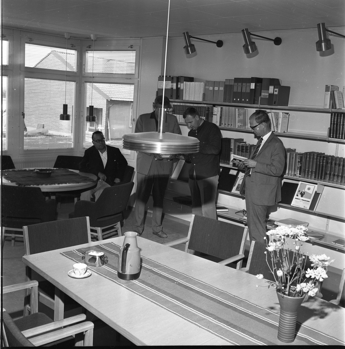 Fyra män i ett lärarrum på Ribbaskolan i Gränna. Ett runt bord med fåtöljer och rektangulärt bord med stolar. Bokhyllor på väggarna med pärmar och böcker. Från vänster: Göte Östlund, Rolf Möller, okänd samt Erik Nyström.