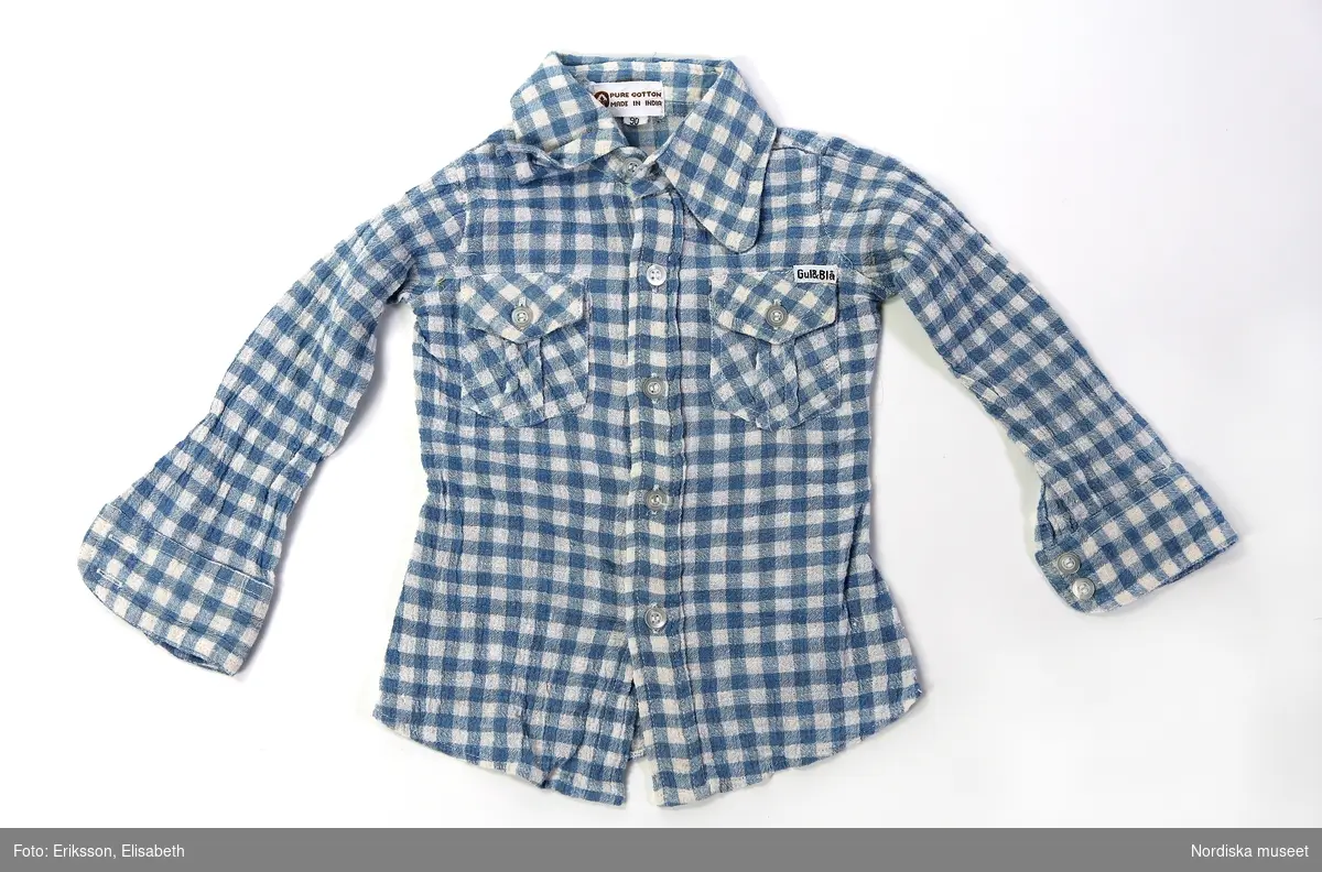 Skjorta från Gul & Blå i barnstorlek 90, rutigt ljusblåvitt crepe yta vävd bomull. 2 fickor framtill med vita knappar. 2 framstycken och 1 bakstycke med ok. 2 insnitt baktill för en figurnära modell. Etikett med texten ”Pure cotton made in India” placerad under kragen på insidan av skjortan. Kragen är nedvikt med höjden 3,5 cm. 5 knappar vid knäppningen och 2 stycken på varje manschett. En liten vit etikett med ”Gul & Blå” i svart text är placerad i ovankant av vänster ficka. Plastfäste för etiketten sitter kvar.

/Maria Vujovic 2018 & Hanna Leijon 2019-03-07