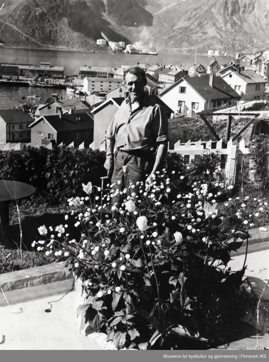 Honningsvåg. Cato Grøtta i hagen utenfor sitt hjem. I bakgrunnen utsikt over deler av bebyggelsen. Ca. 1955.