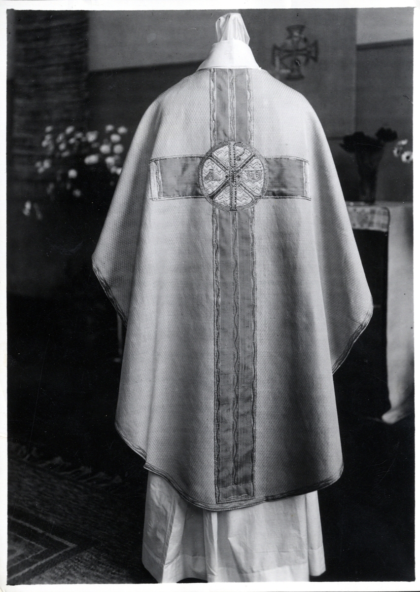 Foto (svart/vitt) av en ljus mässhake (baksidan) med mörkare broderat korsparti, försett med Kristusmonogram. 

Inskrivet i huvudbok 1983.