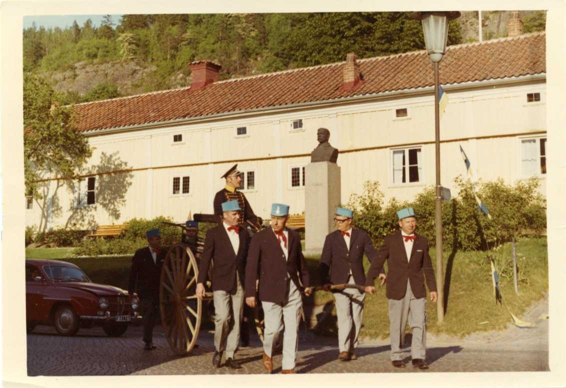 Fem män drar en en-axlad vagn där Olle Mathias sitter klädd i Dolma m/1895 (husaruniform) och modern militär skärmmössa. Männen är klädda i ljus byxor, mörk kavaj med röd fluga samt blå hög mössa på huvudet. Minst tre män går bakom vagnen klädda på samma sätt. Man går på Brahegatan i Gränna och passerar Franckska gården. I bakgrunden till vänster passerar en röd SAAB. I täten går från vänster Per-Olof Forsberg, Elis Moreau, Hans Persson och Nils Lindekrantz
